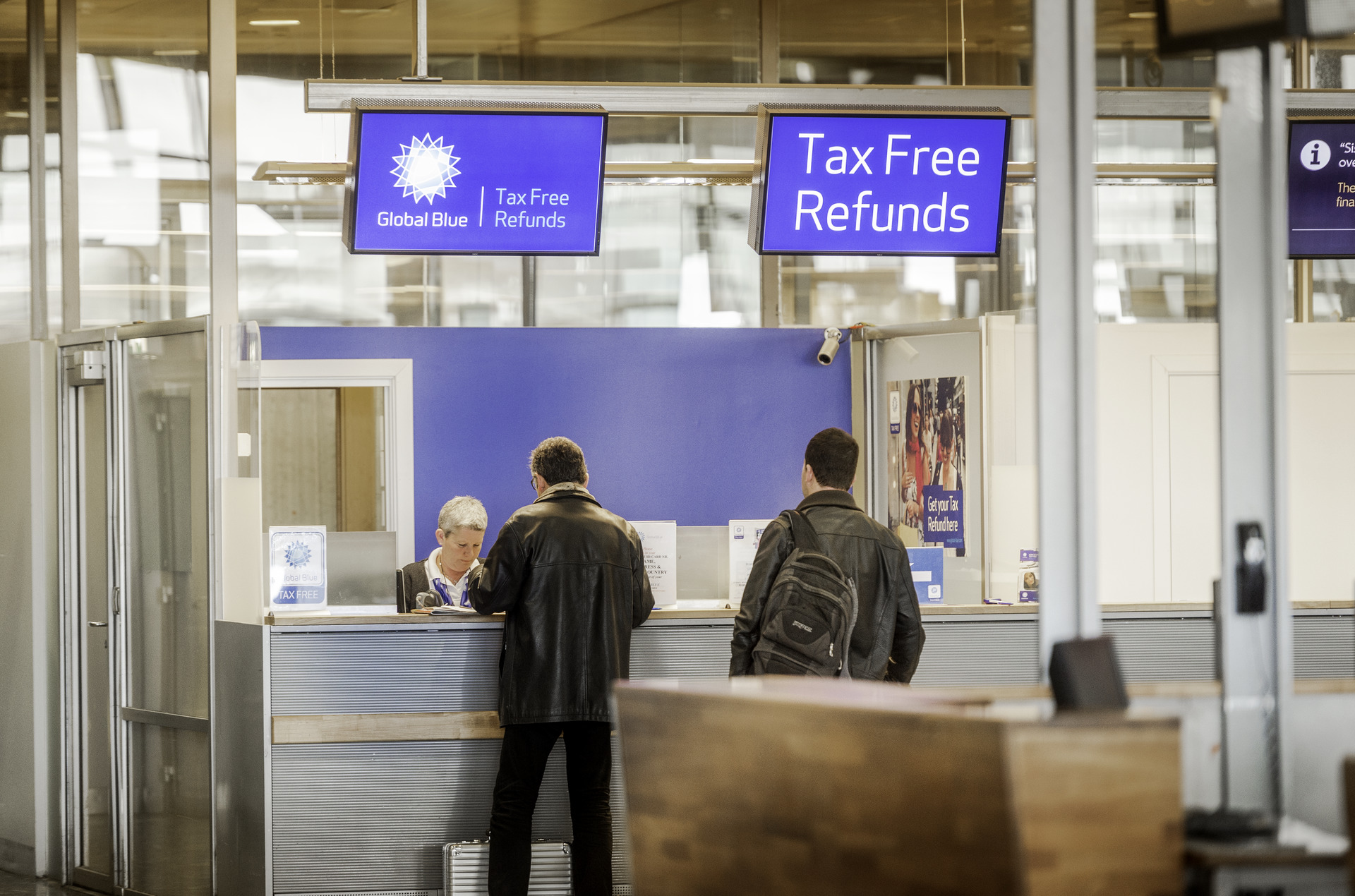 Personer ved en Global Blue tax-free-refusjonsdisk på en flyplass. To menn står ved skranken under et blått skilt som viser "Tax Free Refunds".