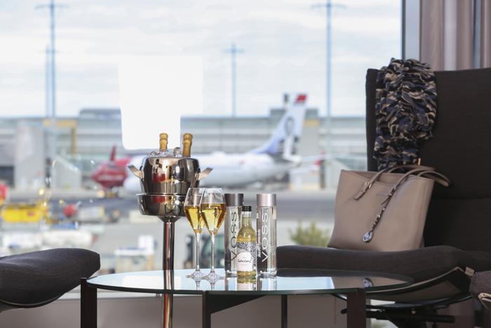 Luksuriøs flyplasslounge med utsikt over rullebane, sjampanje og Voss-flasker på bordet.
